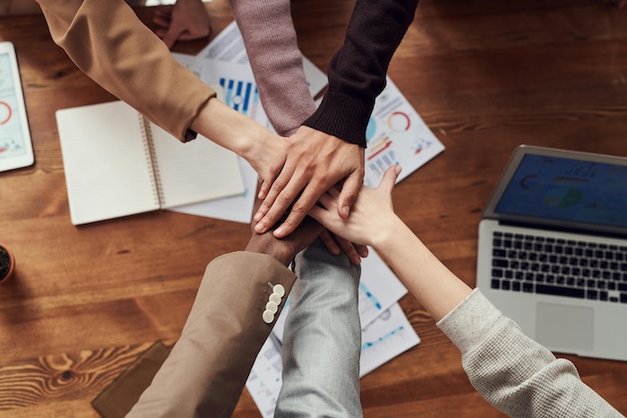 Overhead view of a business team joining hands in unity over a wooden desk with marketing materials, laptop, and business documents scattered beneath. Multiple hands of different skin tones come together in a circle, symbolizing teamwork and collaboration. The scene is captured on a warm wooden surface with various work tools visible in the background.