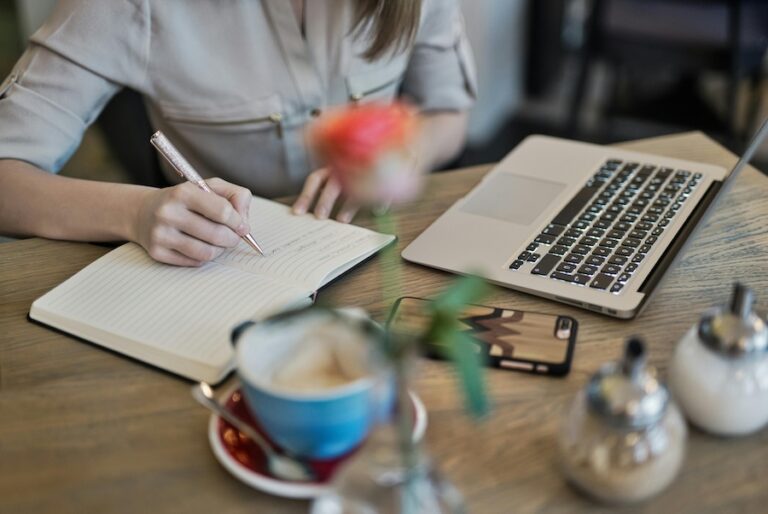 Person writing content marketing strategy for tech companies with a notebook, laptop, and coffee on a desk.